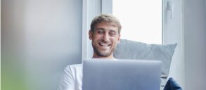 A smiling man looking at his laptop on the couch.