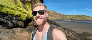 A man smiling to the camera and standing in a rocky coastal area with a gray backpack under a clear blue sky.