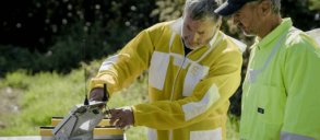 Two people in protective suits outdoors, one demonstrating equipment on a table.