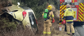 Firefighters responding to a car overturned in a roadside ditch, with a fire truck nearby.