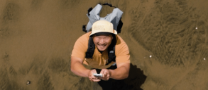 A person wearing a hat and backpack stands on a sandy surface, looking up while holding a smartphone.